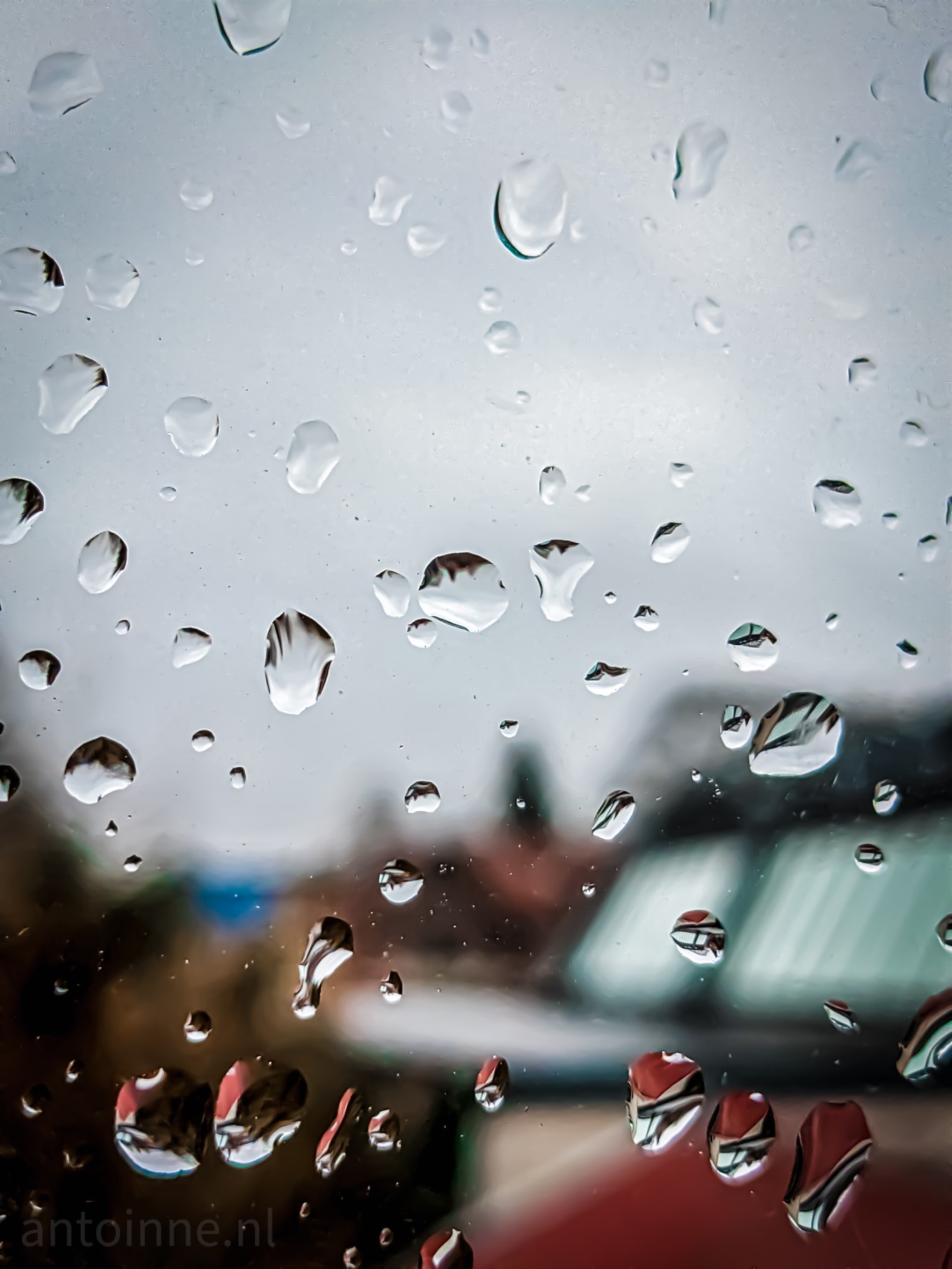 A close-up shot of raindrops on a window. The roofs of other houses are visible in the background. Some are red, others have glass. Far away there is also a burned down building with a blue / green tarpaulin.