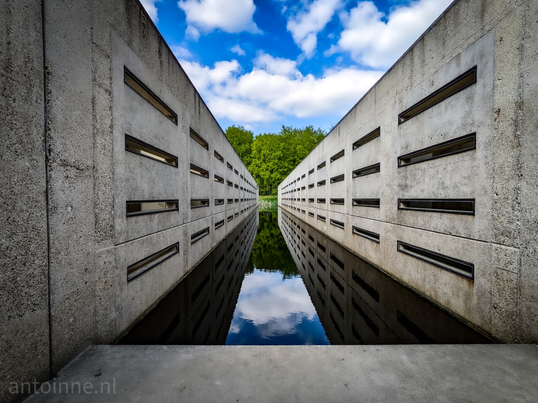 You are standing in the middle of a large monument at a former hydraulic engineering research site. Between the concrete you can see a narrow band of cloudy sky and some trees. These are also reflected in the water.